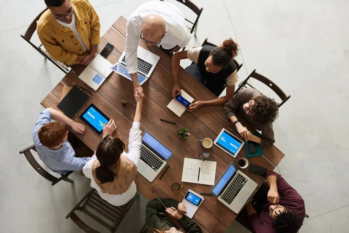Equipo de trabajo alrededor de una mesa con portátiles, visto desde arriba, con dos compañeros dándose la mano