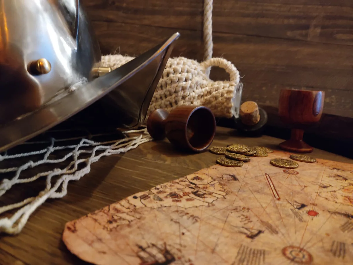 A morion helmet, a net, wooden goblets, gold coins and an old sea chart on a wooden table